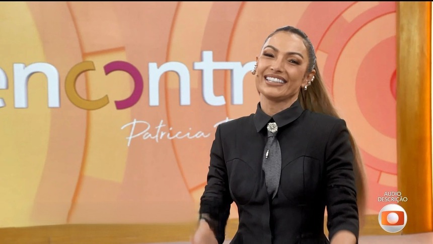 Patricia Poeta com conjunto preto, sorridente, em frente ao telão do programa Encontro