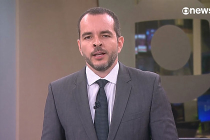Erick Bang, de terno e gravata, em pé no cenário da GloboNews apresentando Plantão Globo