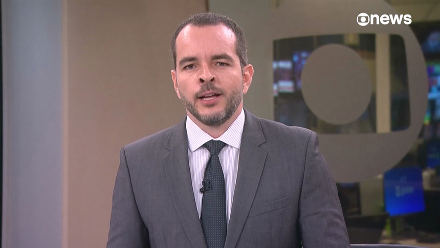 Erick Bang, de terno e gravata, em pé no cenário da GloboNews apresentando Plantão Globo