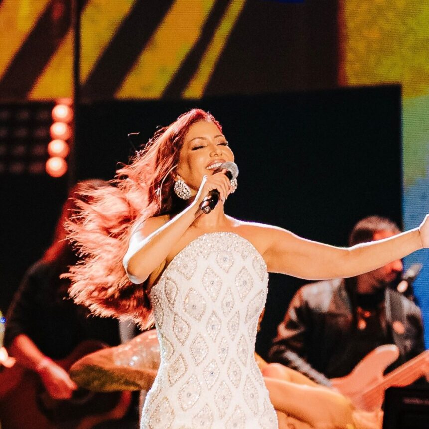 Priscila Senna de vestido branco com brilhantes dourados cantando em cima de um palco