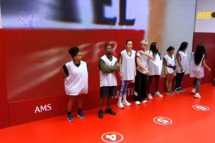 Participantes da Prova do Anjo posicionados em pé em frente ao telão da prova no cenário todo vermelho no BBB 26