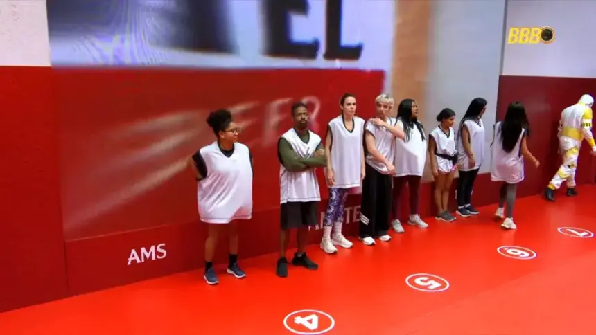 Participantes da Prova do Anjo posicionados em pé em frente ao telão da prova no cenário todo vermelho no BBB 26