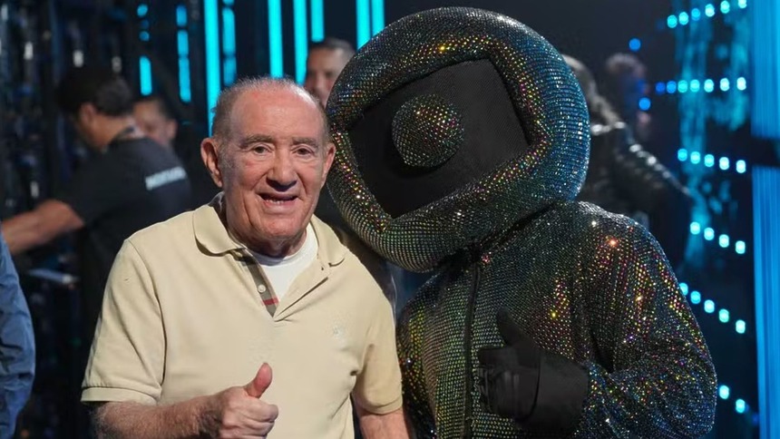 Renato Aragão fazendo o sinal de joia com o dedo, ao lado do mascote com o logo da Globo na cabeça, nos bastidores do Caldeirão