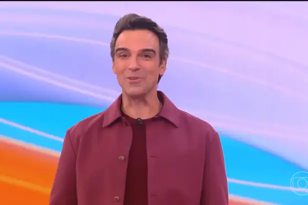 Tadeu Schmidt de blusa vinho sorridente no centro do cenário do BBB 26