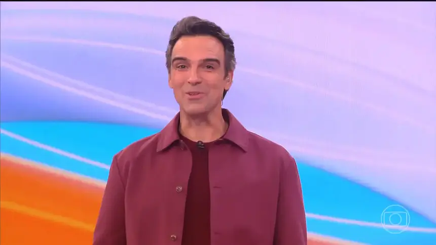 Tadeu Schmidt de blusa vinho sorridente no centro do cenário do BBB 26