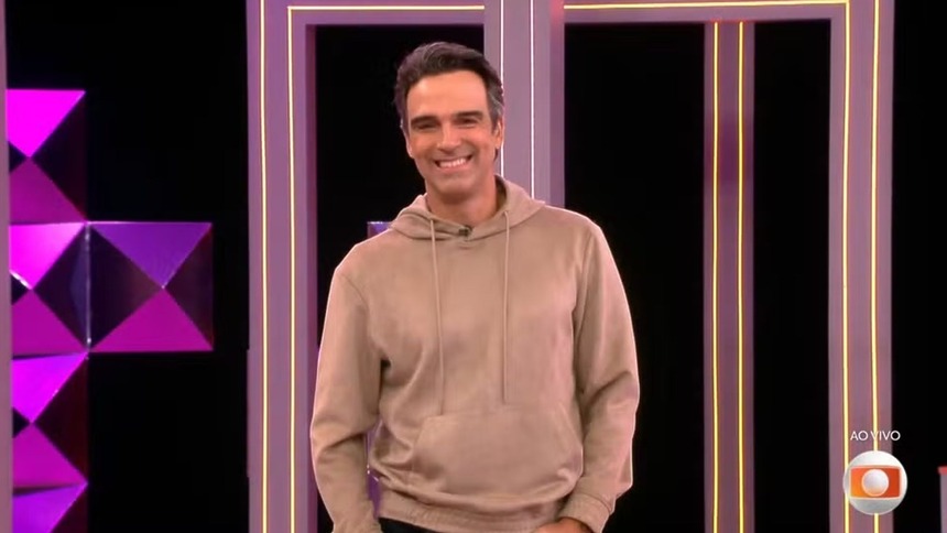 Tadeu Schmidt sorridente, de moletom, falando sobre a Prova do Anjo no BBB 26