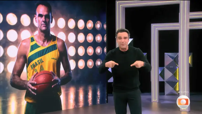 Oscar Schmidt no telão do BBB 26 segurando bola de basquete enquanto Tadeu Schmidt chora em frente ao telão