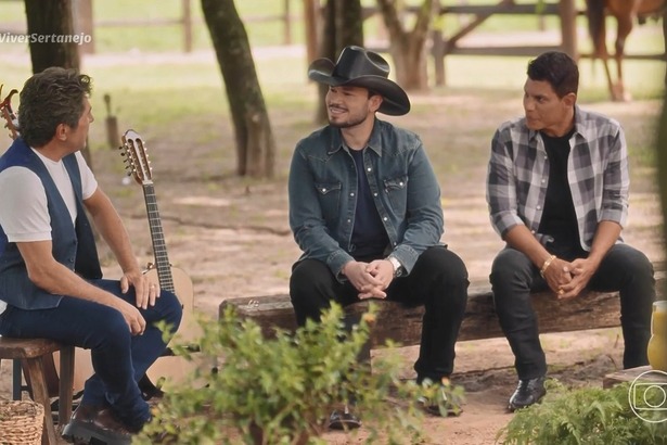 Carreiro & Capataz sentados no banco de madeira olhando para Daniel no Viver Sertanejo