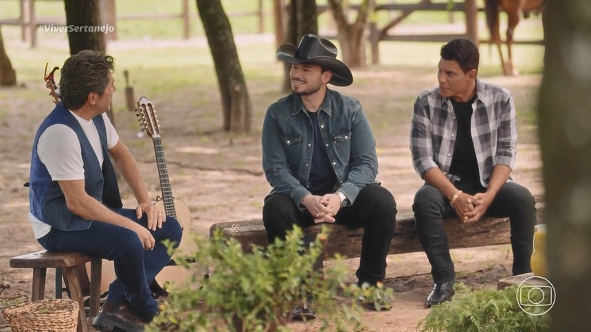 Carreiro & Capataz sentados no banco de madeira olhando para Daniel no Viver Sertanejo