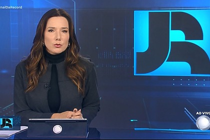 Adriana Perrone veste conjunto preto. Ela está sentada na bancada do Jornal da Record em frente ao telão com o logo do telejornal