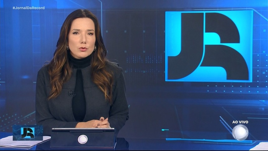 Adriana Perrone veste conjunto preto. Ela está sentada na bancada do Jornal da Record em frente ao telão com o logo do telejornal
