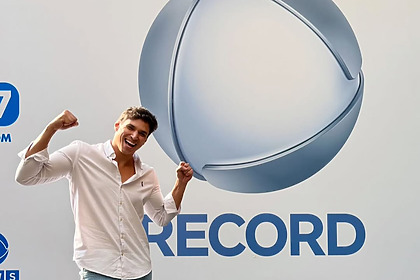 Bruno Cardoso posa sorridente em frente ao logo da Record Entretenimento