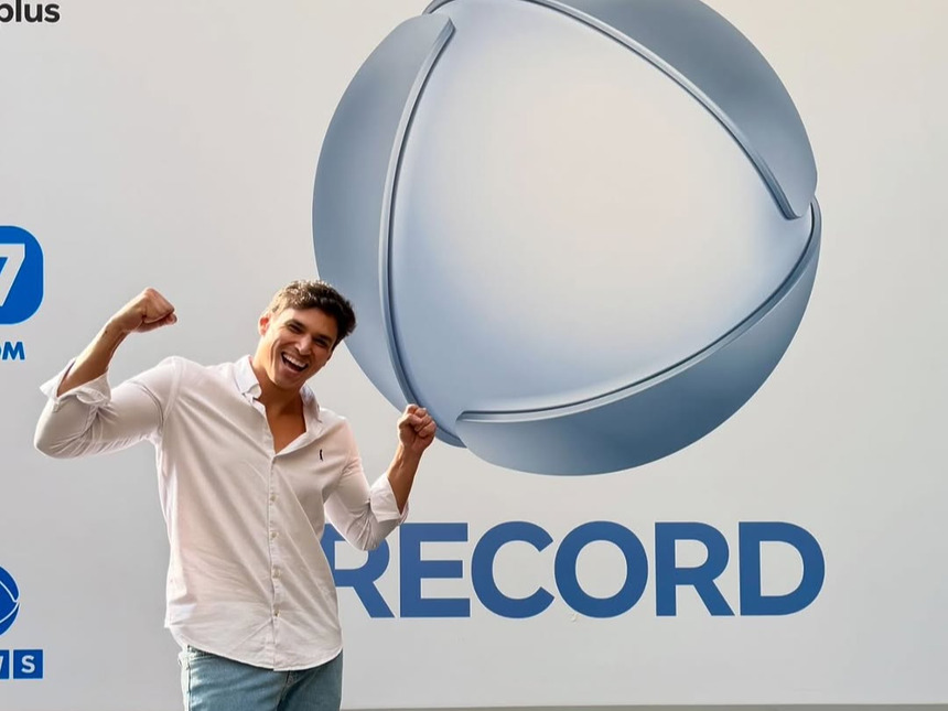 Bruno Cardoso posa sorridente em frente ao logo da Record Entretenimento