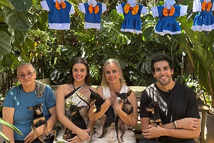 Dona Darci, com os cachorros, a Filha a Neta, ao lado do apresentador Julinho Casares sentados em um banco na reportagem do Hoje Em Dia
