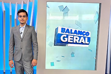 Dudu Camargo de terno cinza em frente a tela vertical do Balanço Geral Manhã. Ele está sorridente