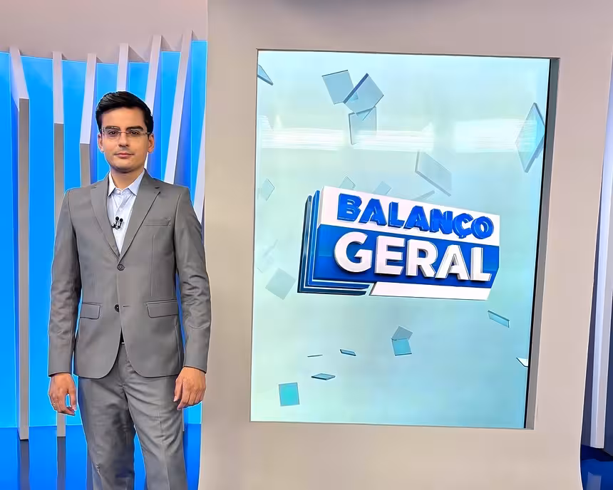 Dudu camargo de terno cinza em frente a tela vertical do balanço geral manhã. Ele está sorridente