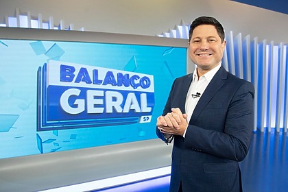 Eleandro Passaia sorridente, em frente ao telão, no cenário do Balanço Geral