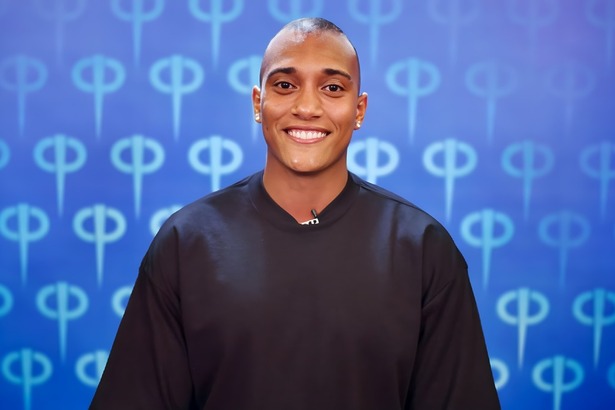 JP sorridente no cenário de apresentação da Casa Do Patrao. Ele veste camisa presta, é um homem moreno com cabelo curto e usa brincos de brilhante