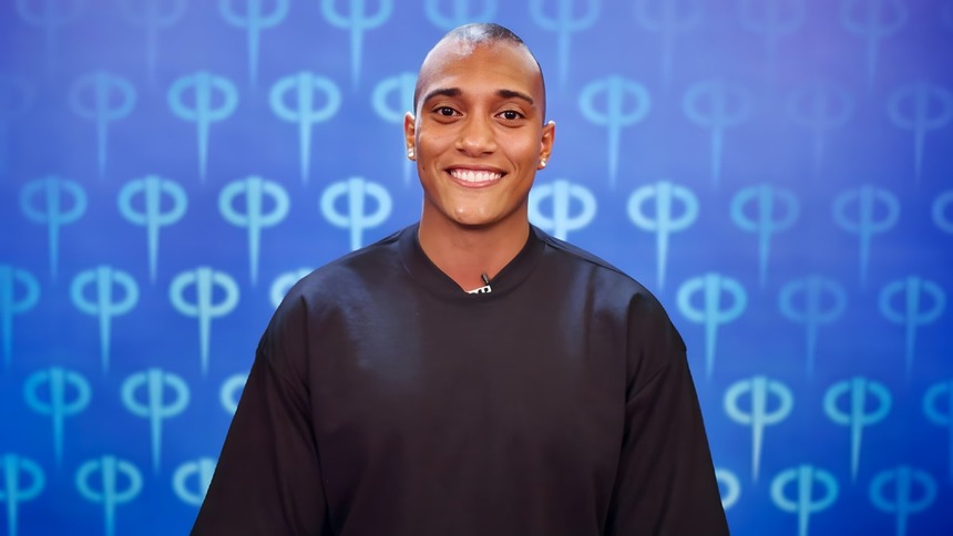 JP sorridente no cenário de apresentação da Casa Do Patrao. Ele veste camisa presta, é um homem moreno com cabelo curto e usa brincos de brilhante