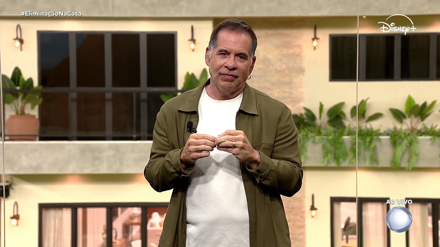 Leandro Hassum de blusa branca e casaco verde, gesticulando, no glass de estúdio da Casa do Patrão