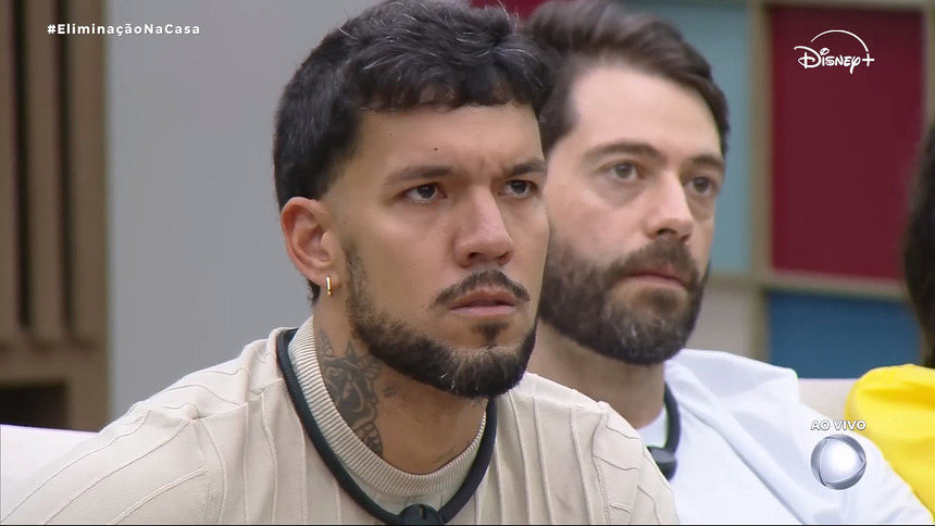 Luis Fellipe sentado no sofá da Casa Do Patrão atento ao telão onde Leandro Hassum discursa para o eliminado