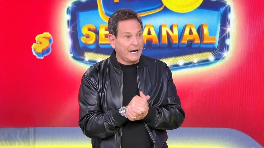 Luis Ricardo caminha no cenário do sorteio da Tele Sena Semanal. O cenário está com artes gráficas na cor vermelha da edição 105