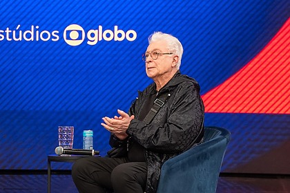 Aguinaldo Silva sentado em uma poltrona em frente a um telão com o logo dos Estúdios Globo
