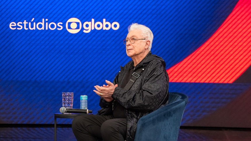 Aguinaldo Silva sentado em uma poltrona em frente a um telão com o logo dos Estúdios Globo