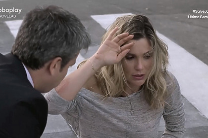 Erica (Flávia Alessandra) sentada no chão de uma avenida com a mão na cabeça desesperada sendo acolhida por Haroldo (Otaviano Costa) em cena de Salve Jorge