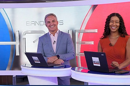 Leandro Stoliar e Thalyta Almeida sorridentes na bancada do BandNews Hoje. Eles estão em pé frente ao telão com o logo do telejornal nas cores azul e vermelho