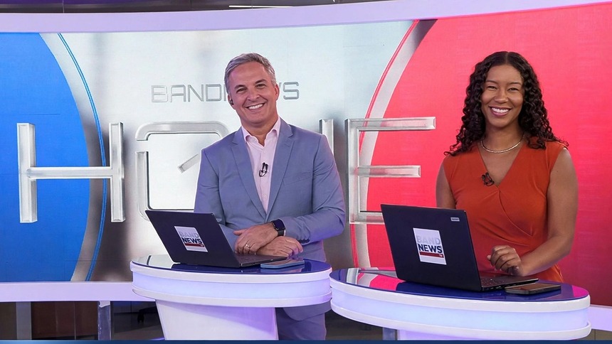 Leandro Stoliar e Thalyta Almeida sorridentes na bancada do BandNews Hoje. Eles estão em pé frente ao telão com o logo do telejornal nas cores azul e vermelho