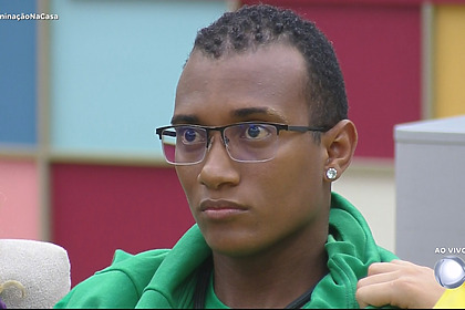 JP de camisa verde olha sério para o telão da sala da Casa do Patrão após Leandro Hassum anunciar sua eliminação