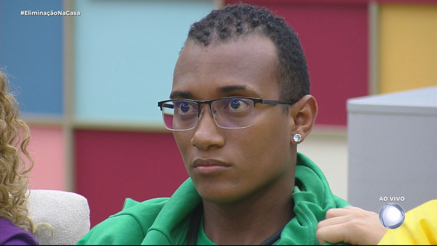 JP de camisa verde olha sério para o telão da sala da Casa do Patrão após Leandro Hassum anunciar sua eliminação