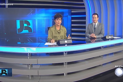 Christina Lemos e Eduardo Ribeiro sentados na bancada do Jornal da Record sendo seguidos por uma câmera em movimento
