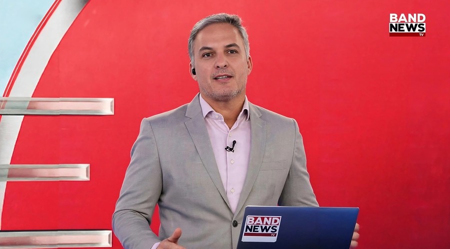 Leandro Stoliar em frente ao telão na cor vermelha e com o notebook aberto no novo cenário do BandNews TV