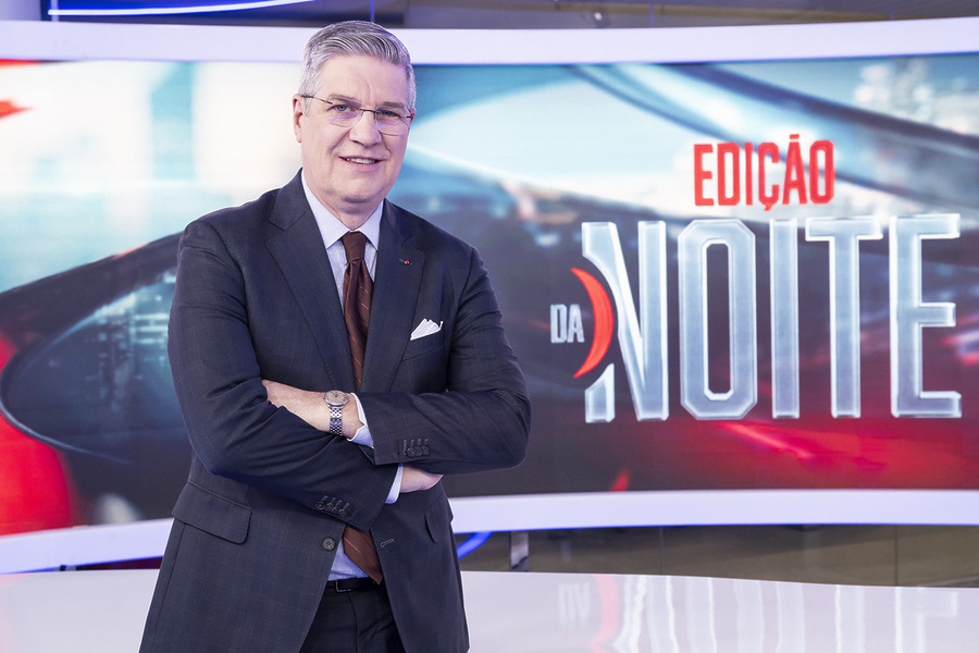 Felipe Vieira sorridente, em pé,  na frente da bancada do Edição da Noite