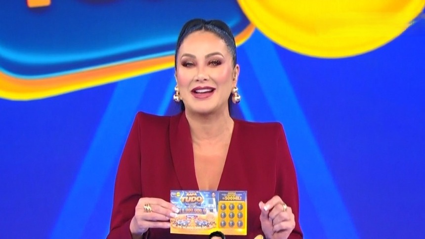 Helen Ganzarolli, veste um conjunto vermelho e segura o bilhete no palco do terceiro Sorteio da Tele Sena De Pais 2026