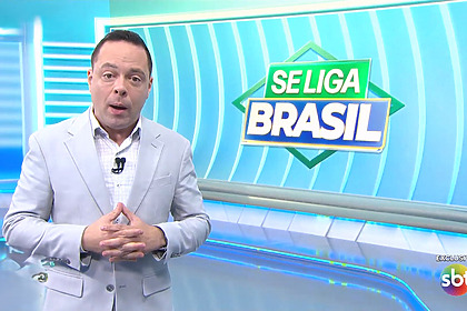 Thiago Gardinali em pé de terno claro no cenário do Se Liga Brasil do SBT