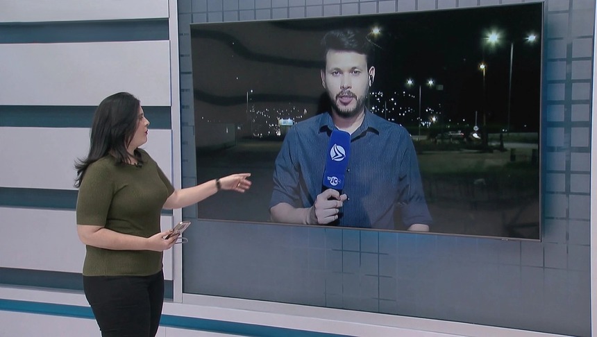 Apresentadora Suelen Brainer, no estúdio da TV Jornal, aponta para o telão onde está o repórter Jean Ferreira segurando o novo microfone azul do SBT
