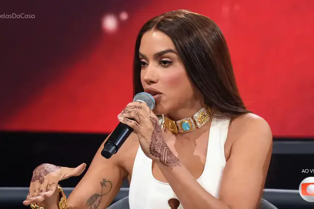 Anitta fala no microfone do Estrelas da Casa. Ela está sentada e veste uma roupa branca