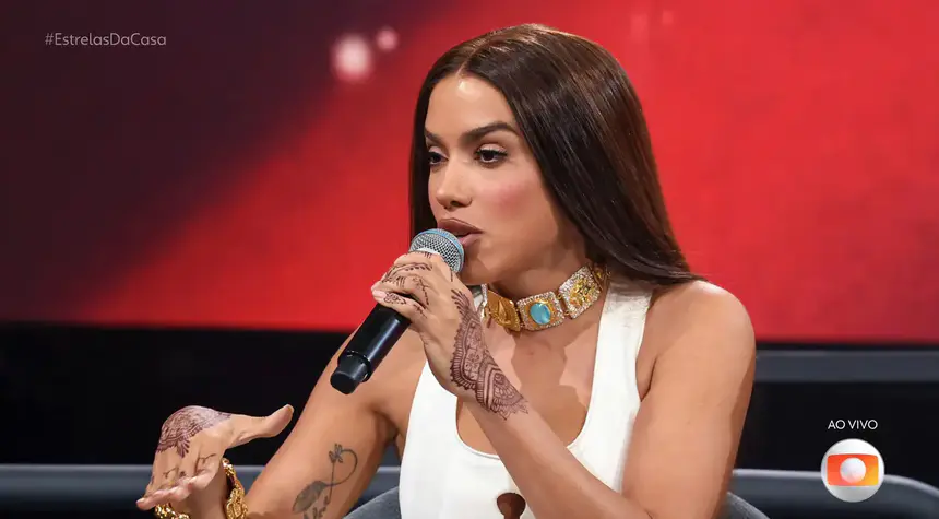 Anitta fala no microfone do Estrelas da Casa. Ela está sentada e veste uma roupa branca