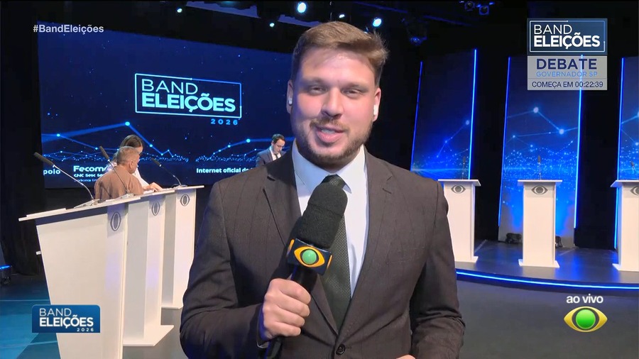 Repórter Pedro Teixeira segura microfone da Band e sorrir no centro do cenário do Debate na Band
