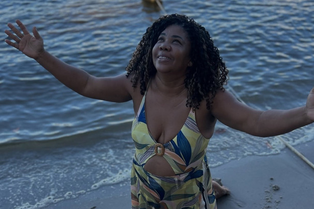 Mãe De Davi Brito ajoelhada na areia de uma praia com as mãos para o céu