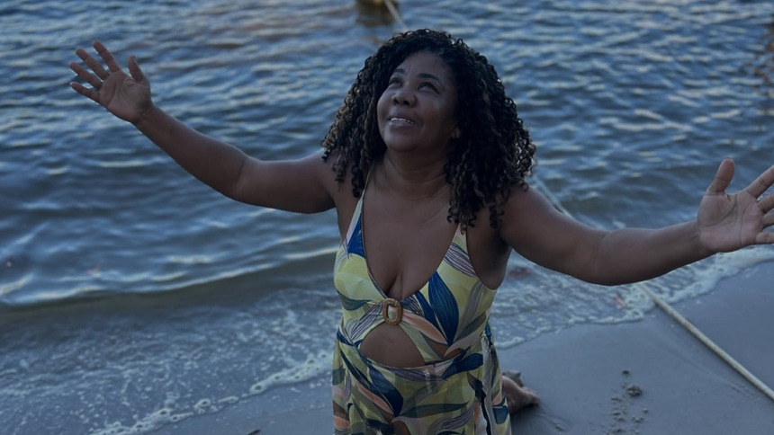 Mãe De Davi Brito ajoelhada na areia de uma praia com as mãos para o céu