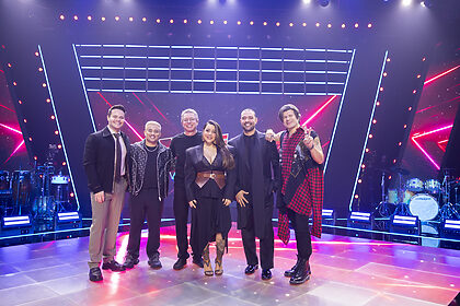 The Voice Brasil Treinadores Com Boninho em pé no centro do palco do reality show