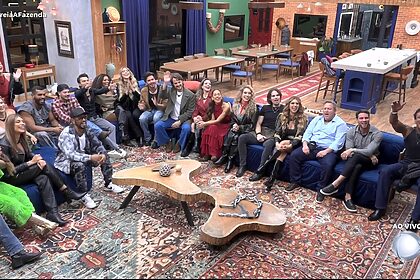 Elenco completo de A Fazenda 18 sentados na sala olhando para o telão onde está Adriane Galisteu