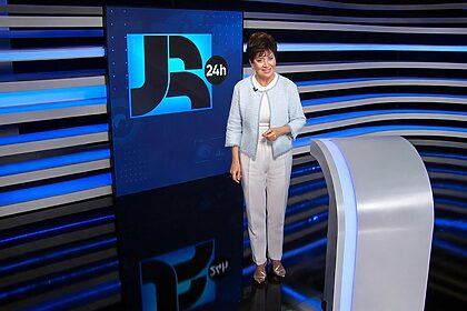 Christina Lemos em pé e sorrindo no encerramento do JR 24h. Ela veste roupas claras