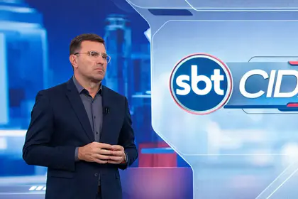 Rodrigo Bocardi com semblante sério em pé na frente do logo do SBT Cidades