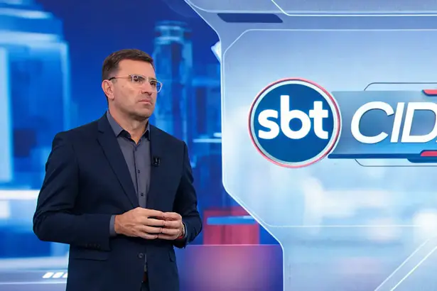 Rodrigo Bocardi com semblante sério em pé na frente do logo do SBT Cidades