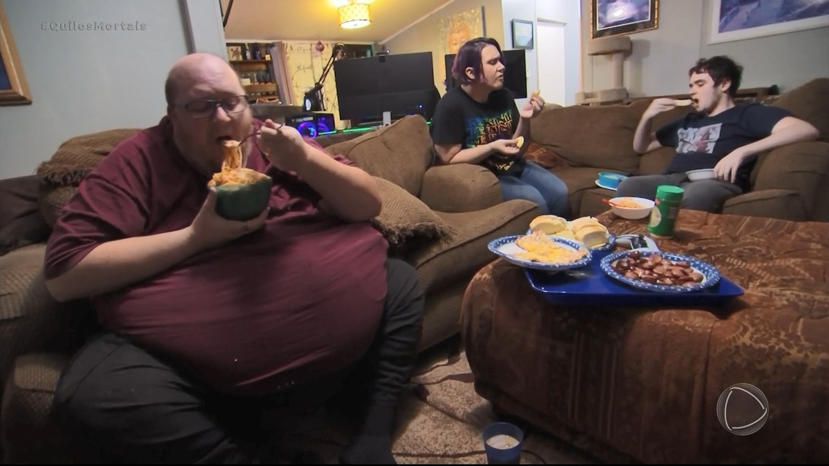 Michael comendo macarrão sentado no sofá com uma barriga bem acentuada